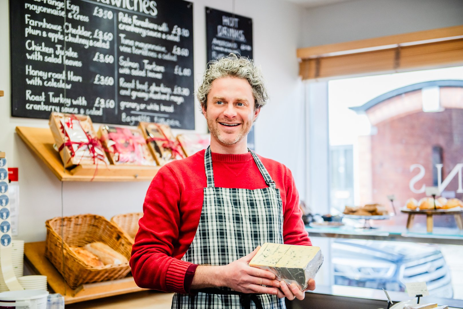 man in red jumper and checked apron holding half a wheel of cheese at Saxtons deli in Church Stretton